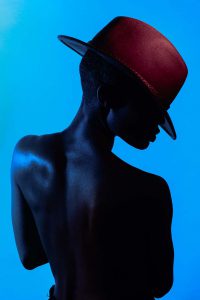 Capture the sultry, mysterious side of a beautiful woman as she stands shirtless wearing a hat, illuminated by dynamic lighting. The play of light and shadow highlights her curves and creates an air of intrigue. Discover the art of seduction with this captivating photograph.