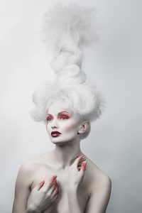 In this stunning high-fashion photography, a nude model with porcelain white hair and touches of red makeup is portrayed, wearing a dramatic long hairpiece. The contrast between the model's pale complexion and the bold red accents creates a striking and eye-catching image. The composition is artistic and sophisticated, showcasing the model's body and features in a tasteful and elegant way. With this captivating image, the photographer skillfully captures the essence of contemporary fashion, while highlighting the model's unique beauty and individuality.