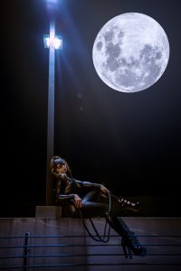 This image features Catwoman sitting seductively under a street light with the moon above her, creating an alluring and mysterious atmosphere. The black and white composition of the photo, paired with the dramatic lighting, gives it a noir-like quality that adds to the overall allure.