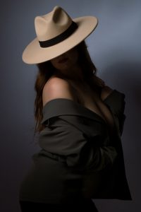 A striking image of a sultry woman with a bold fashion sense, dressed in a stunning blazer and hat, captured under dramatic lighting with deep shadows. The play of light and shadow creates a mysterious and alluring vibe that draws the viewer in, making it impossible to look away. This image is a testament to the photographer's artistry in creating visually captivating portraits that leave a lasting impression.